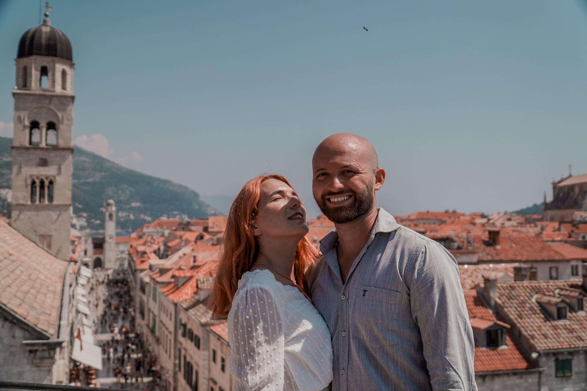 A couple is standing on the old walls of Dubrovnik, enjoying their journey with smiles on their faces. Behind them, you can see the old town with its red roofs, the clock tower, and the main street.