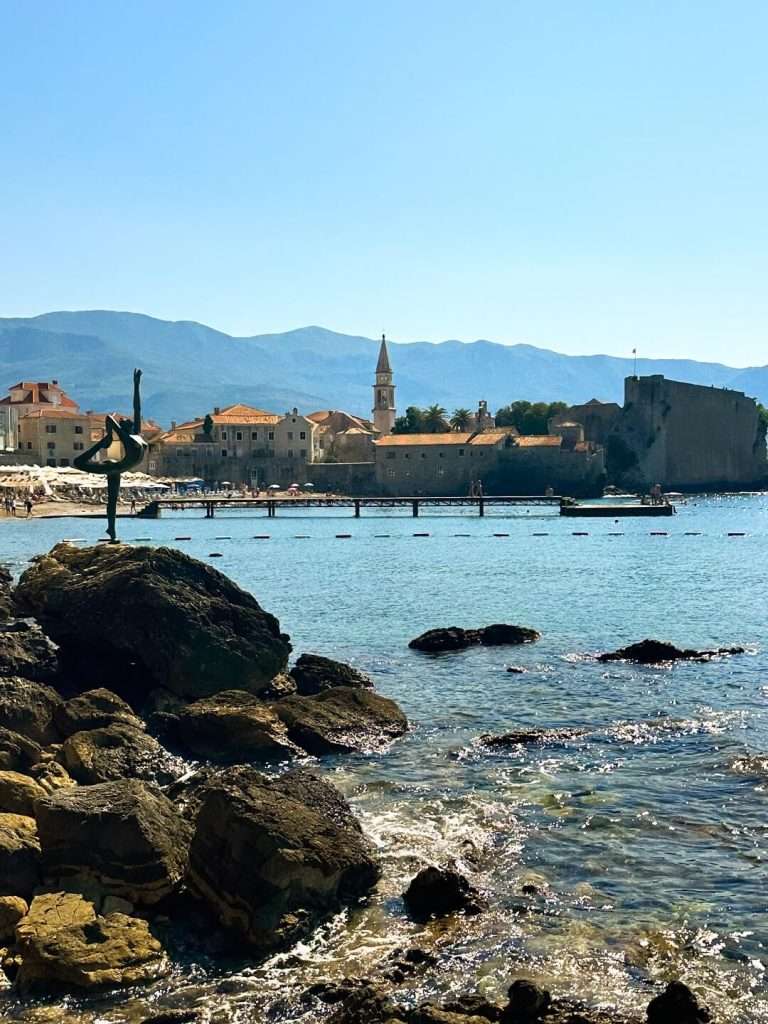 At the edge of the sea, there are rocks with a statue of a ballerina dancing on top. Behind the sea, you can see the old town of Budva with its stone buildings and walls.