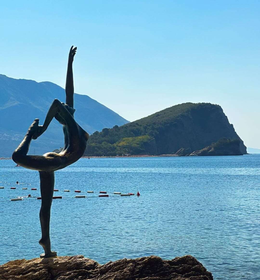 A bronze statue of a ballerina standing on a rock. Behind her, you can see the Adriatic Sea and the island of Sveti Nikola.